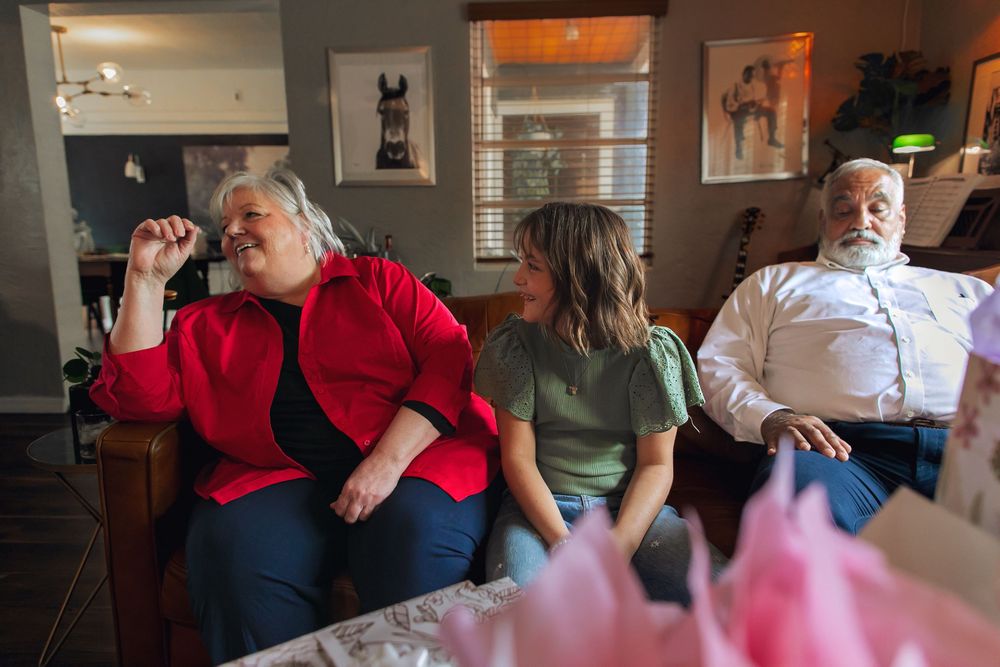 Three people sitting on a couch in a living room with pink decorations visible in the foreground.