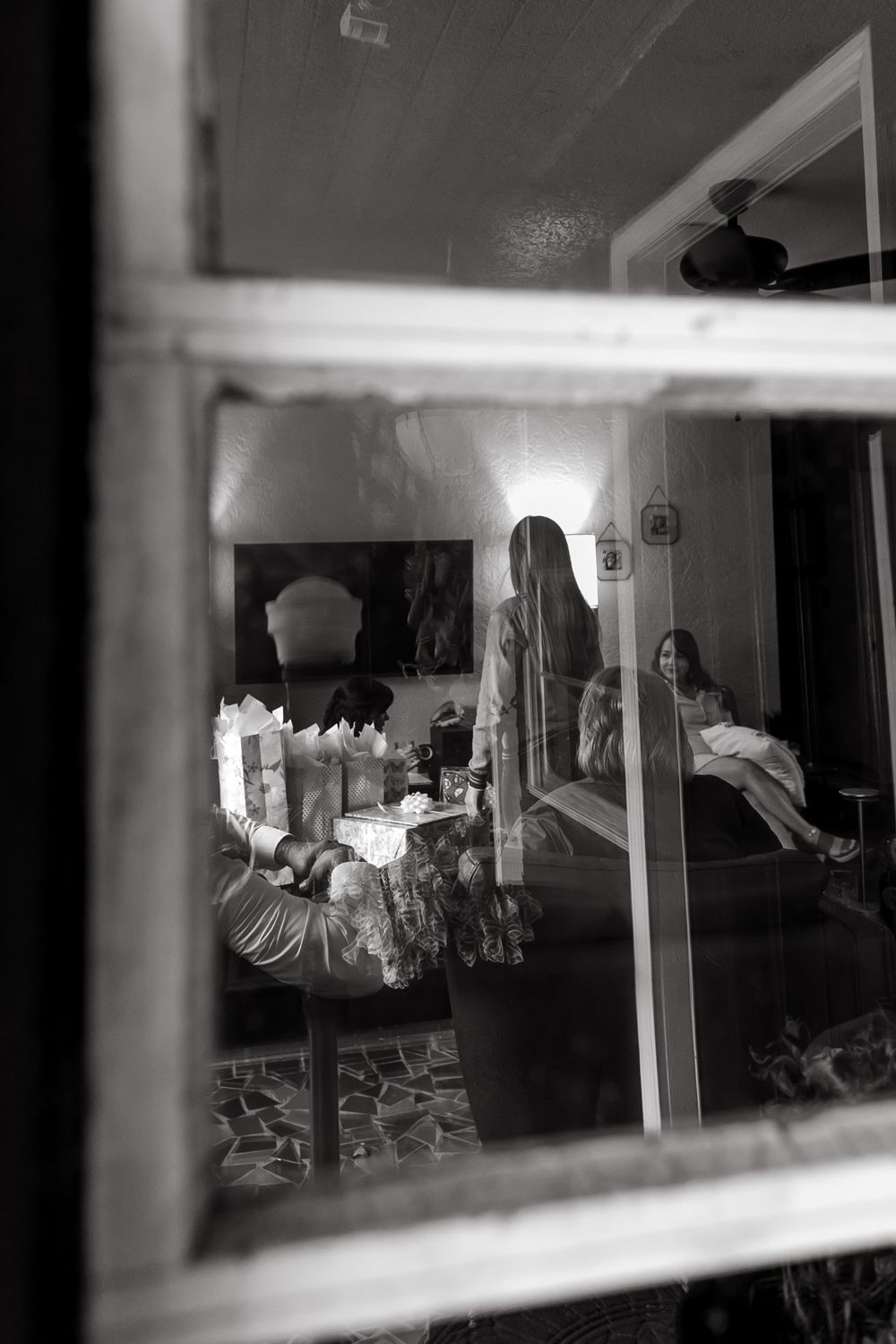 A moody black and white photo looking through a window into a dimly lit room with a mirror reflection.