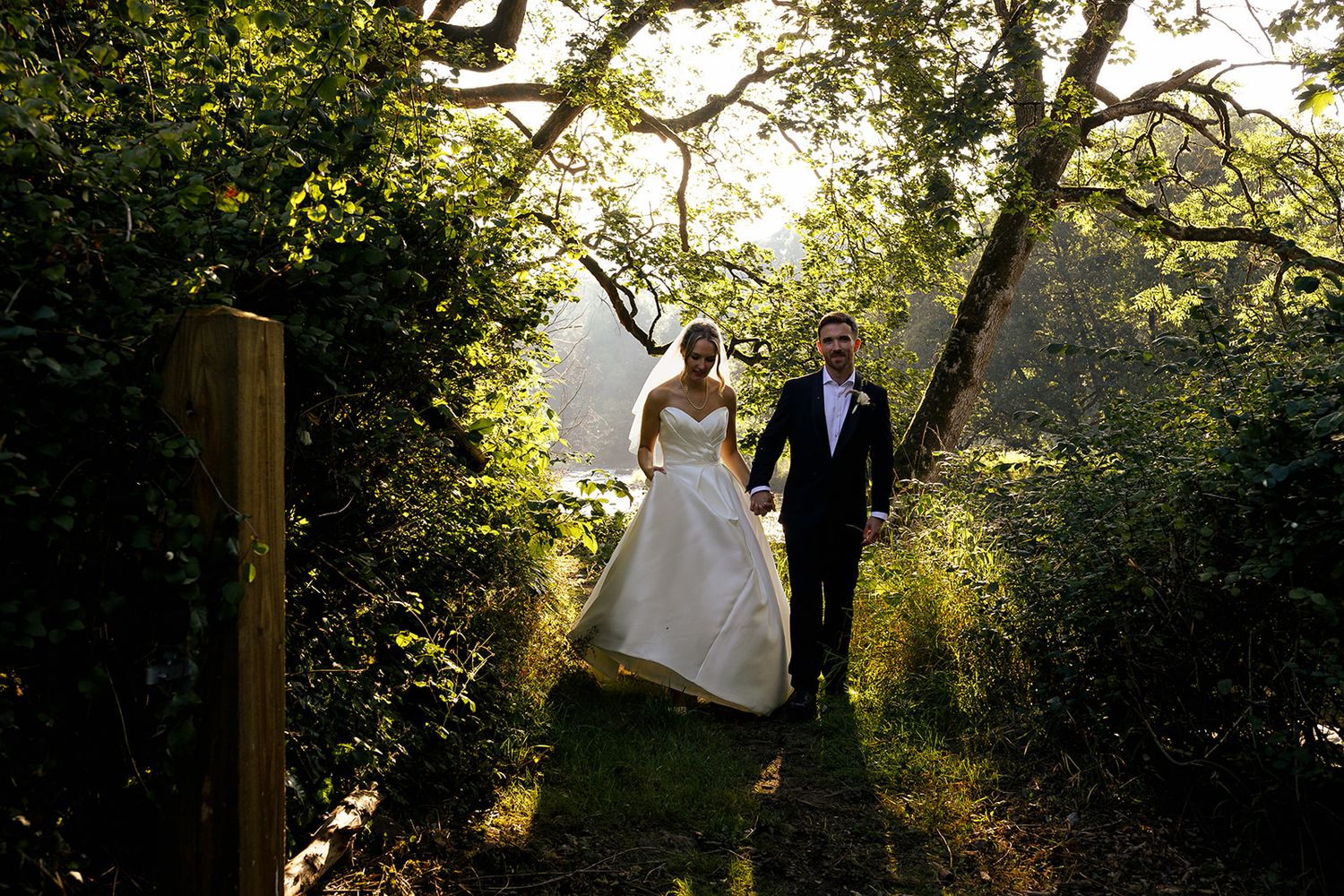 A bride and groom walk hand-in-hand down a path surrounded by lush greenery and trees.
