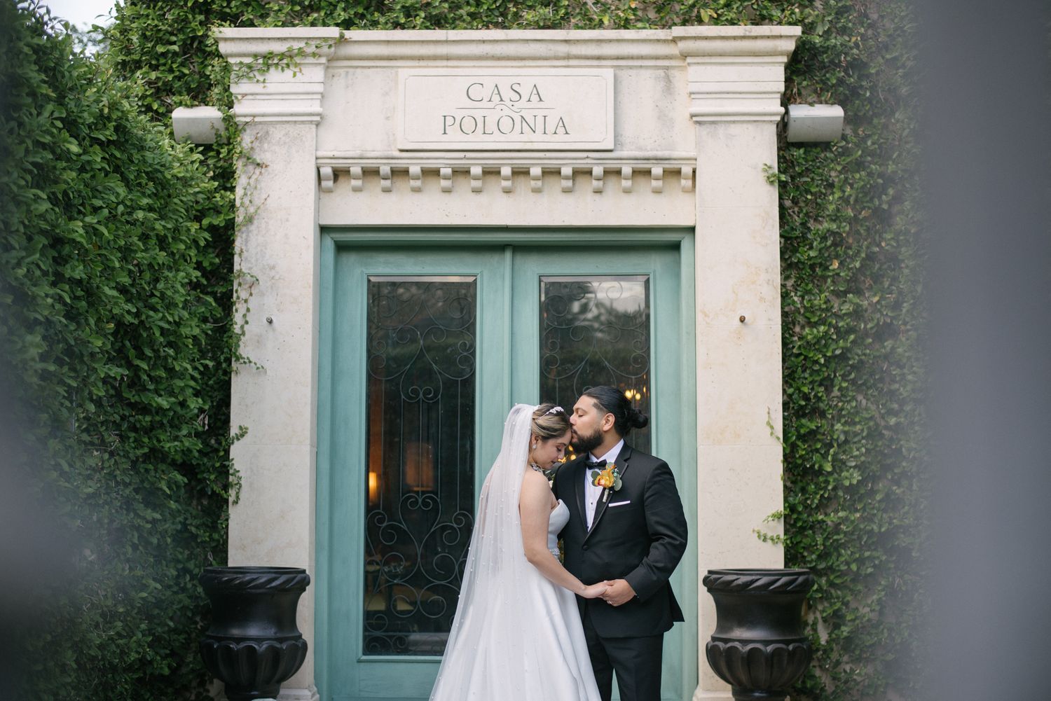 A couple in wedding attire embraces in front of an elegant white stone building entrance with blue-green doors.