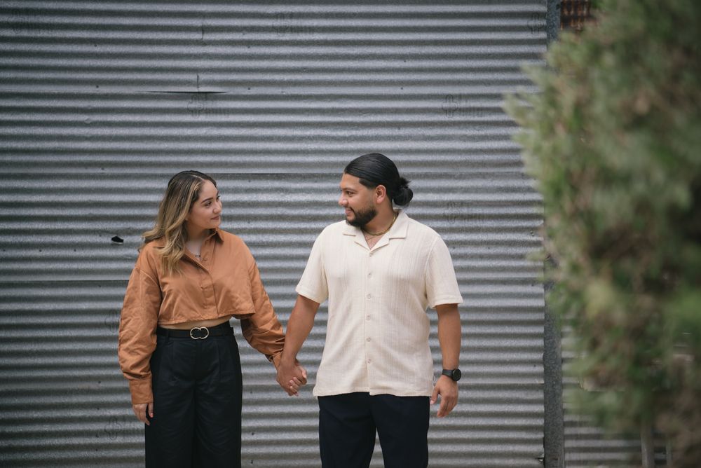 A couple stands together holding hands and smiling in front of an industrial metal wall.