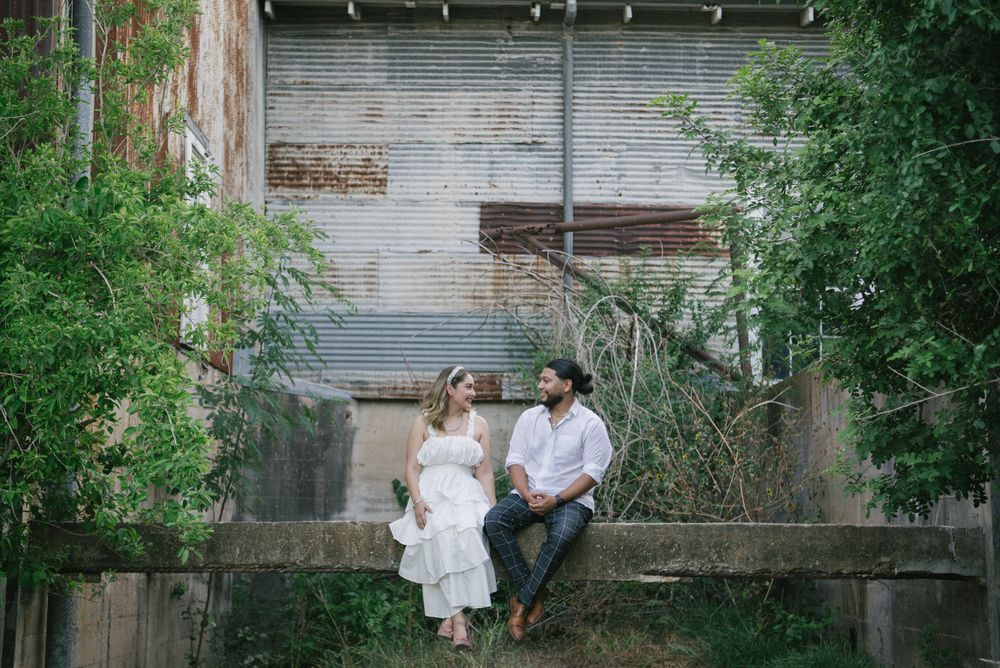 A couple sits together on a rustic wooden fence surrounded by lush greenery and a brick wall background.
