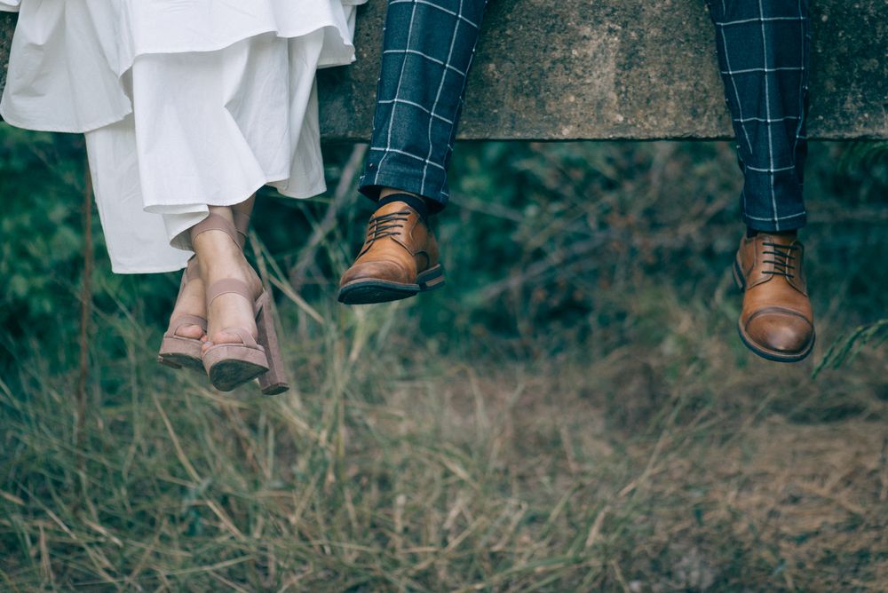 Two pairs of feet in brown leather shoes dangle together over an earthy background with dry grass.