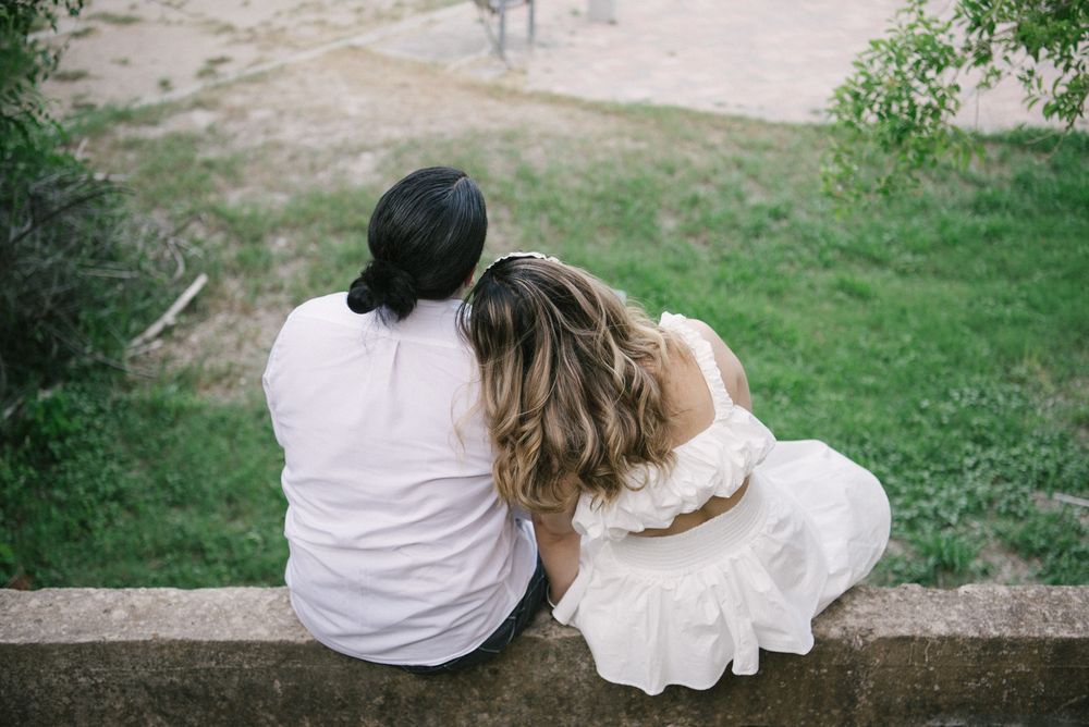 Two people in light colored clothing sit close together on concrete ledge overlooking grassy area.