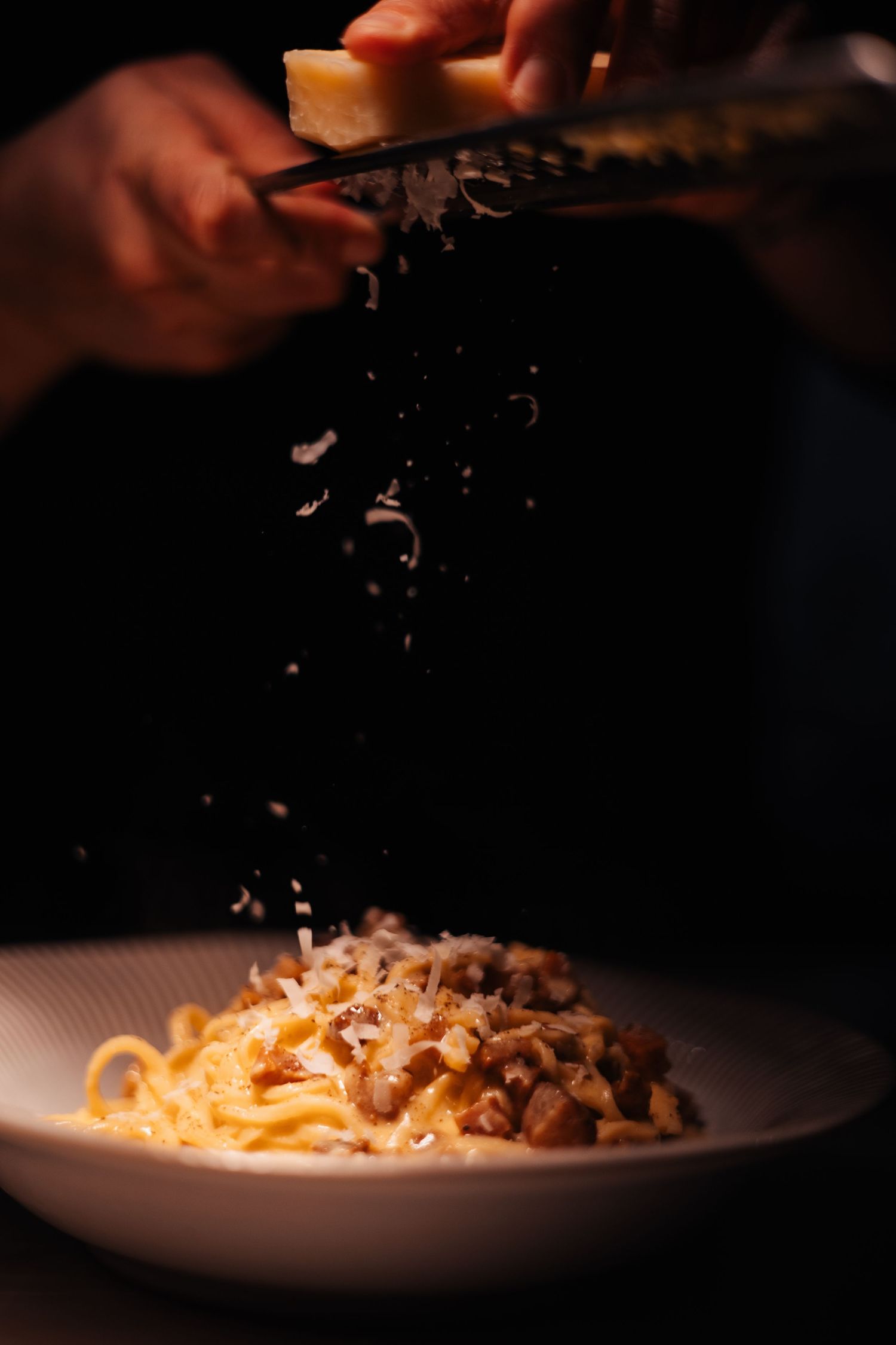 Fresh pasta being garnished with grated cheese in dramatic lighting.