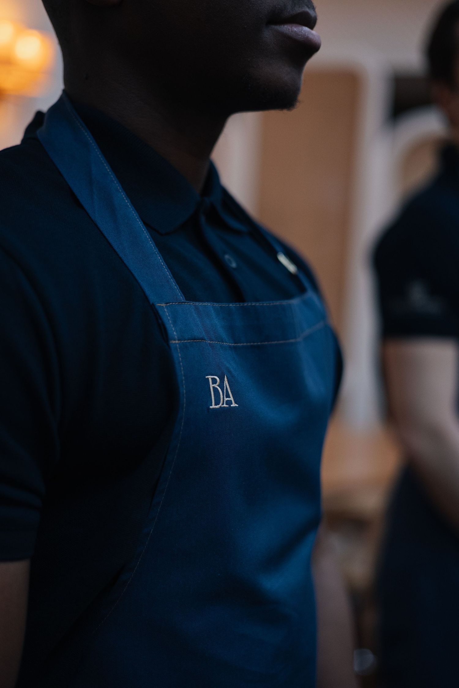 Restaurant staff wearing navy blue aprons with embroidered lettering stand in a row.