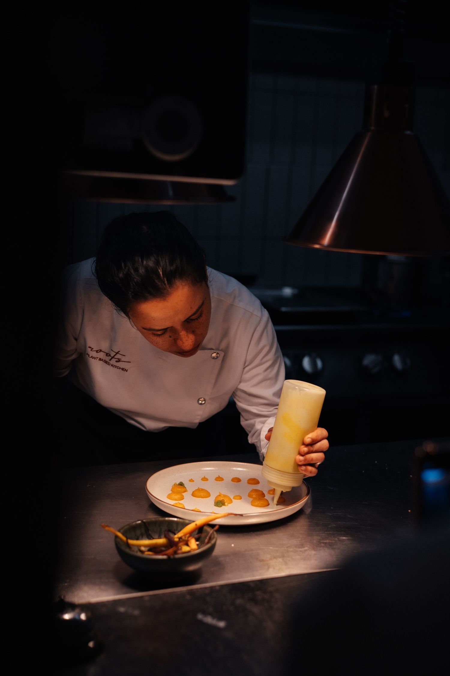 Chef in white uniform carefully inspecting and plating dishes in a dimly lit kitchen.