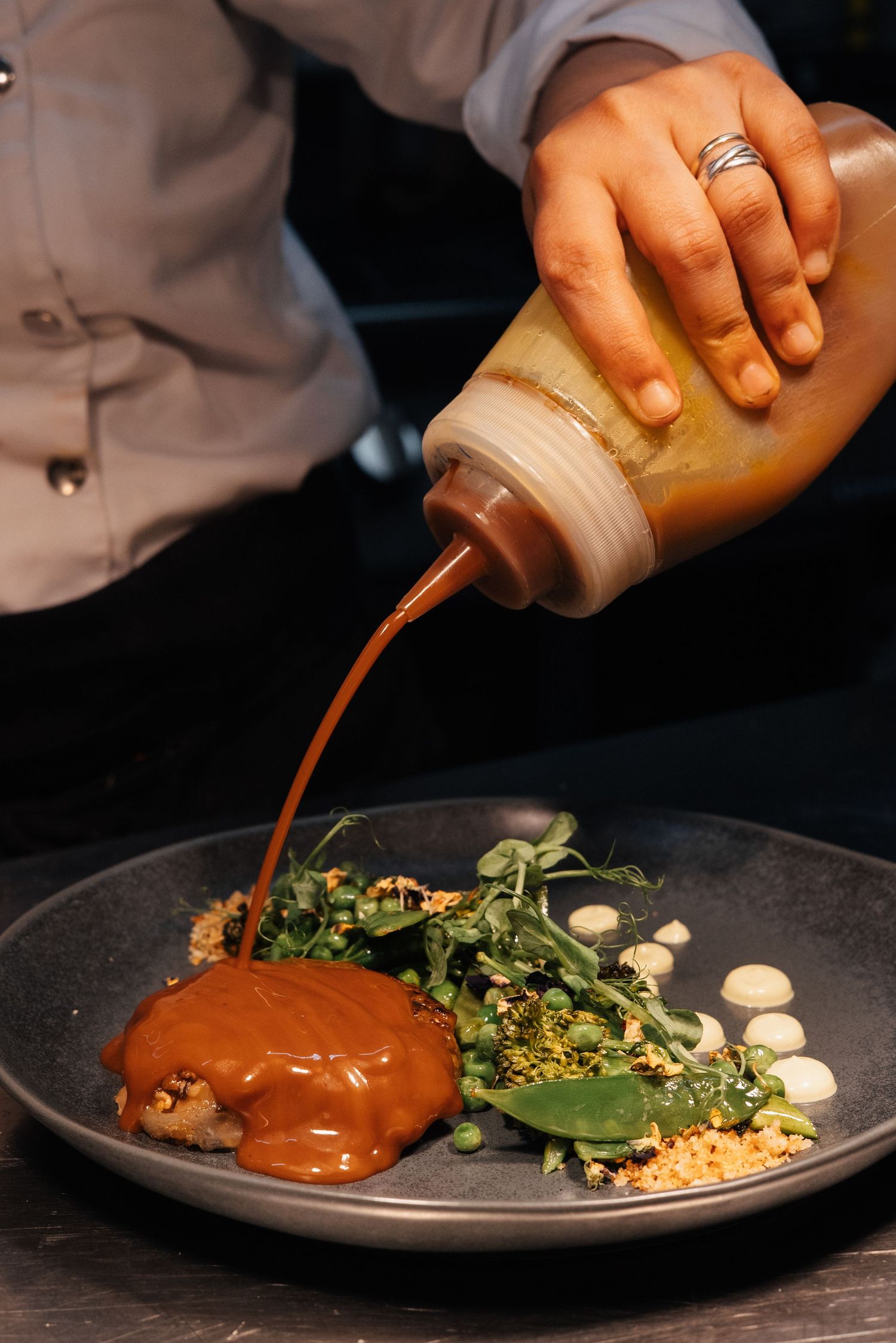 Hands pouring rich brown sauce over a carefully plated dish with greens and garnishes.