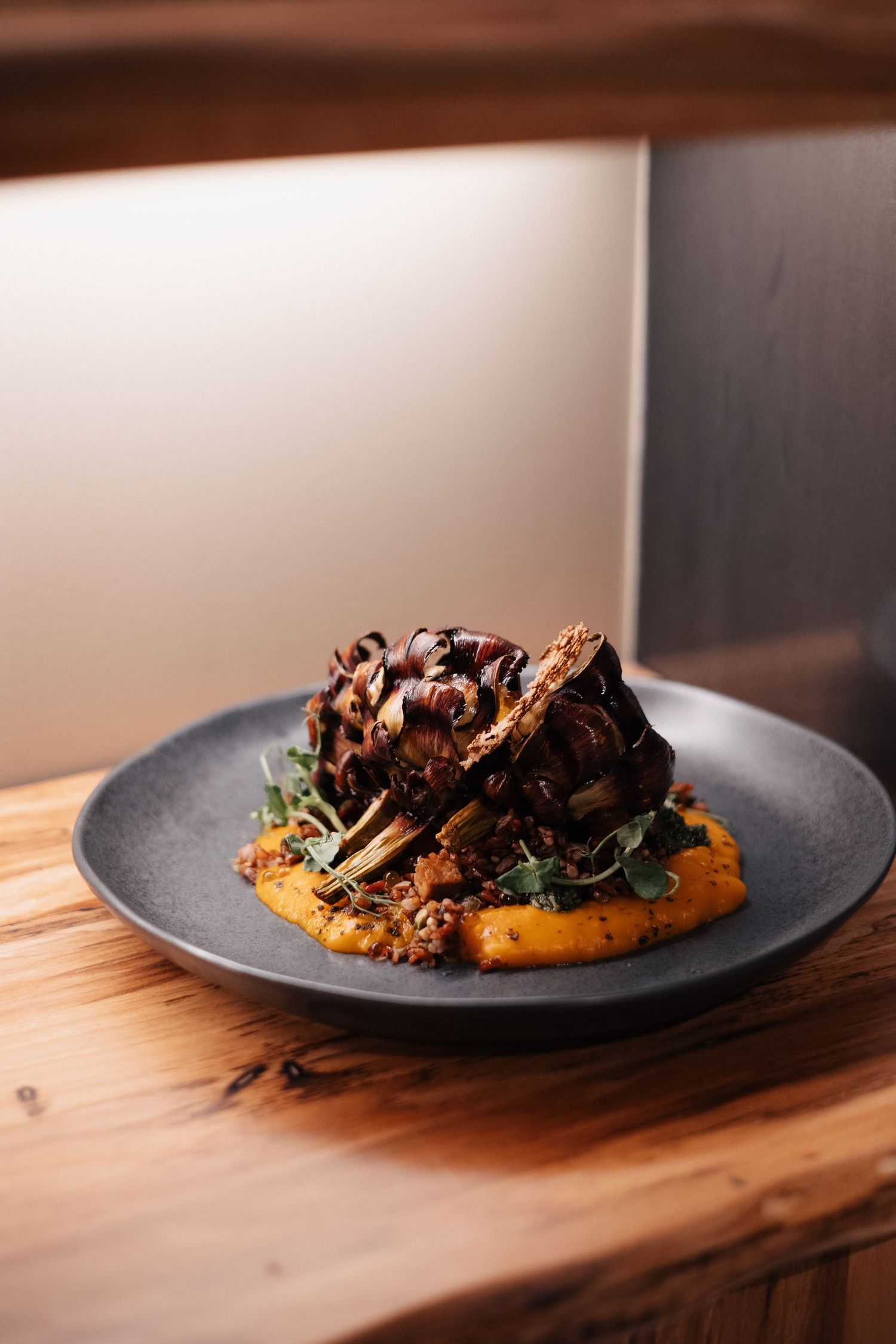 Gourmet meat dish served on black plate with orange puree and fresh herbs on wooden table.