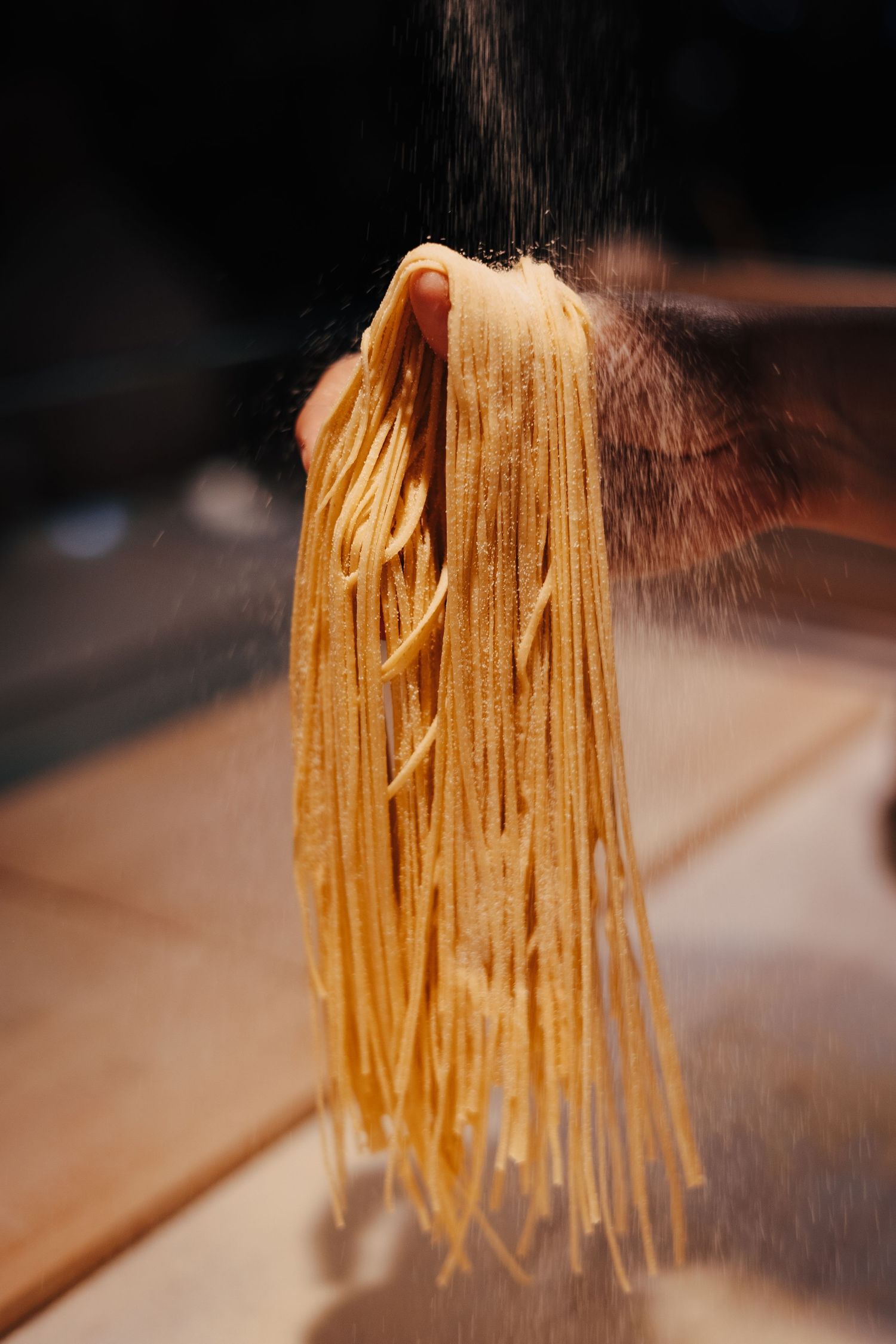 Fresh homemade pasta strands being lifted against dark background with flour dusting.