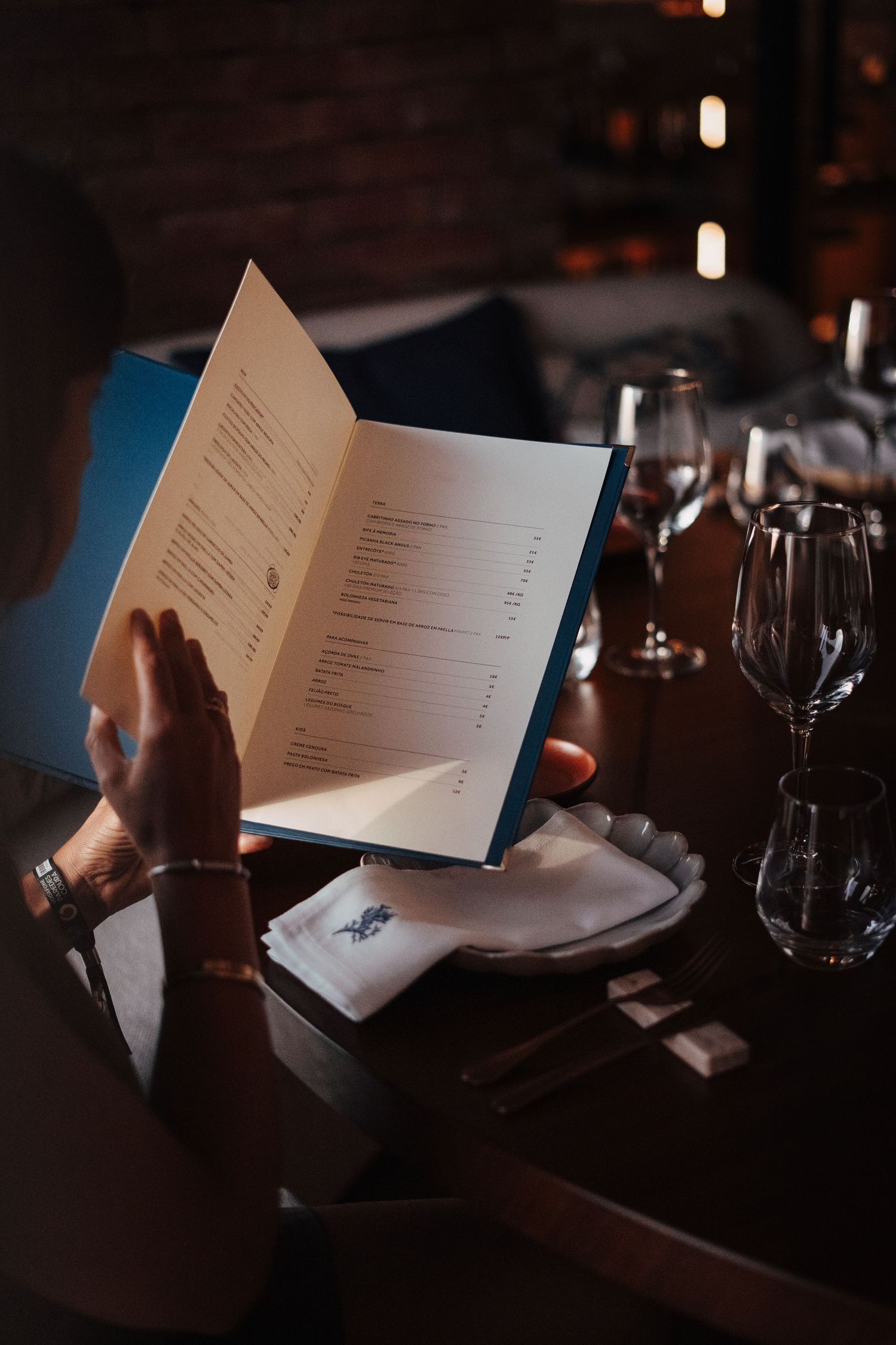Hands holding restaurant menu in dimly lit dining setting with wine glasses.