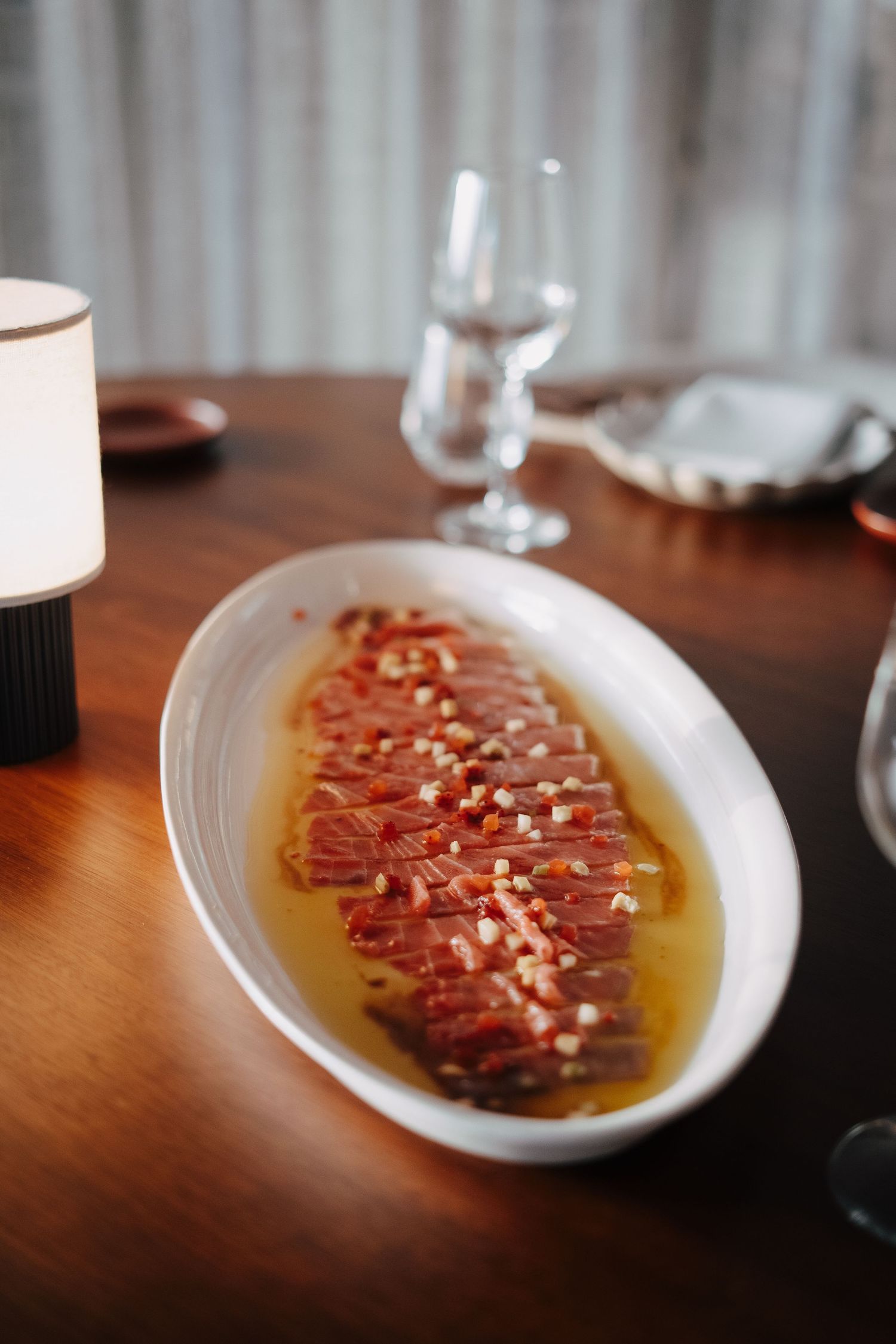 Thinly sliced prosciutto drizzled with olive oil and garnished with pine nuts on a white plate.