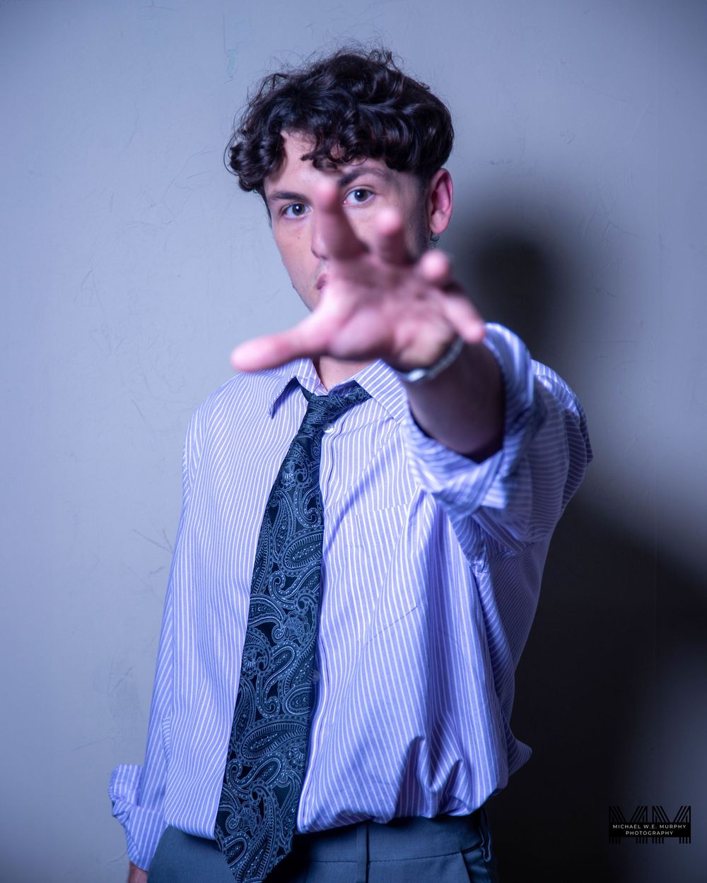 Person in striped dress shirt and tie pointing directly at camera with finger gun gesture in dramatic lighting.