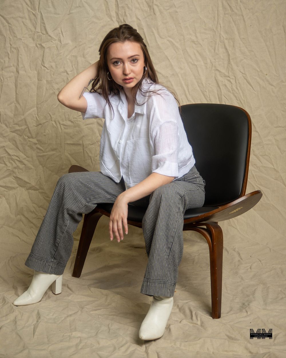 Studio portrait of someone in a white button-down shirt and gray jeans sitting casually on a modern wooden chair.