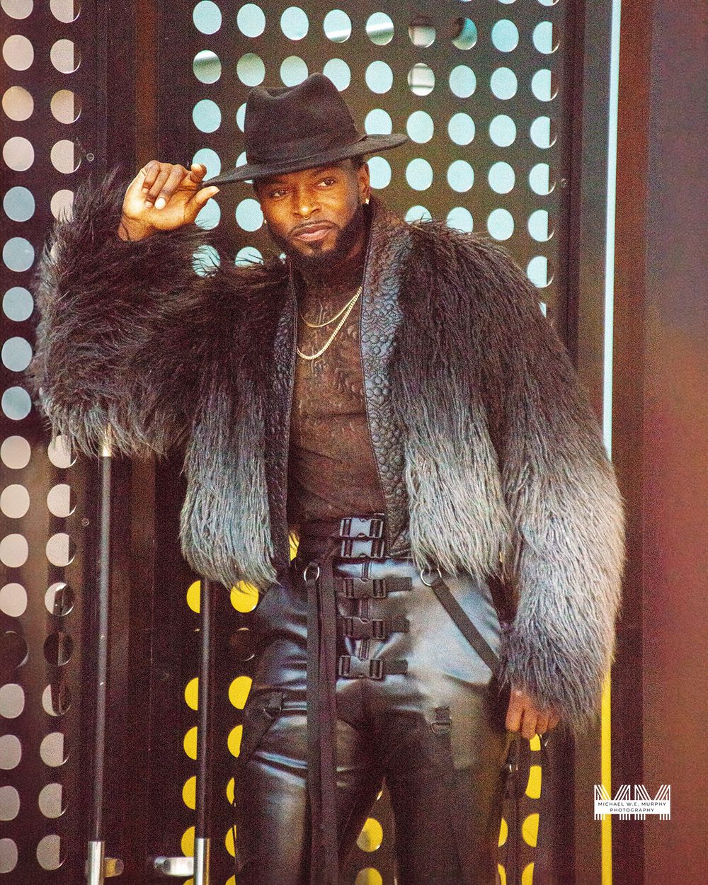 Performer in black fur coat, brown shirt and leather pants poses in front of illuminated polka dot backdrop.