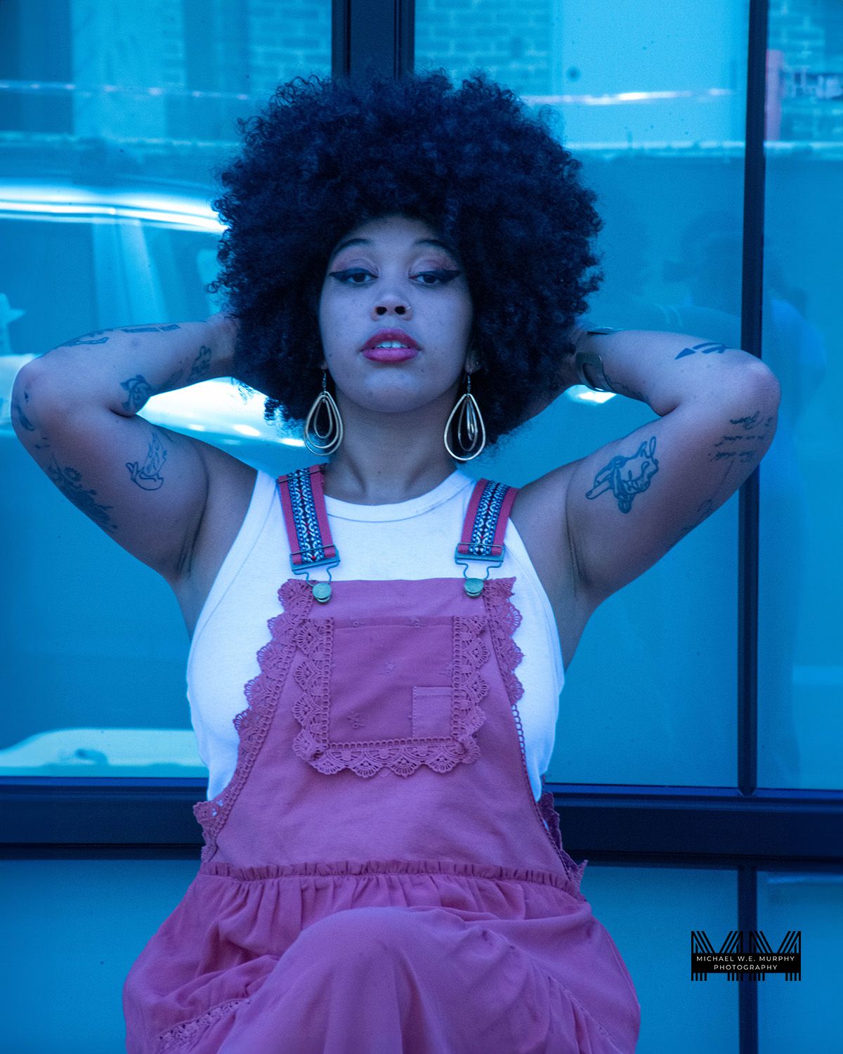 A retro fashion portrait with a dramatic afro hairstyle and pink overalls against a blue-tinted background.