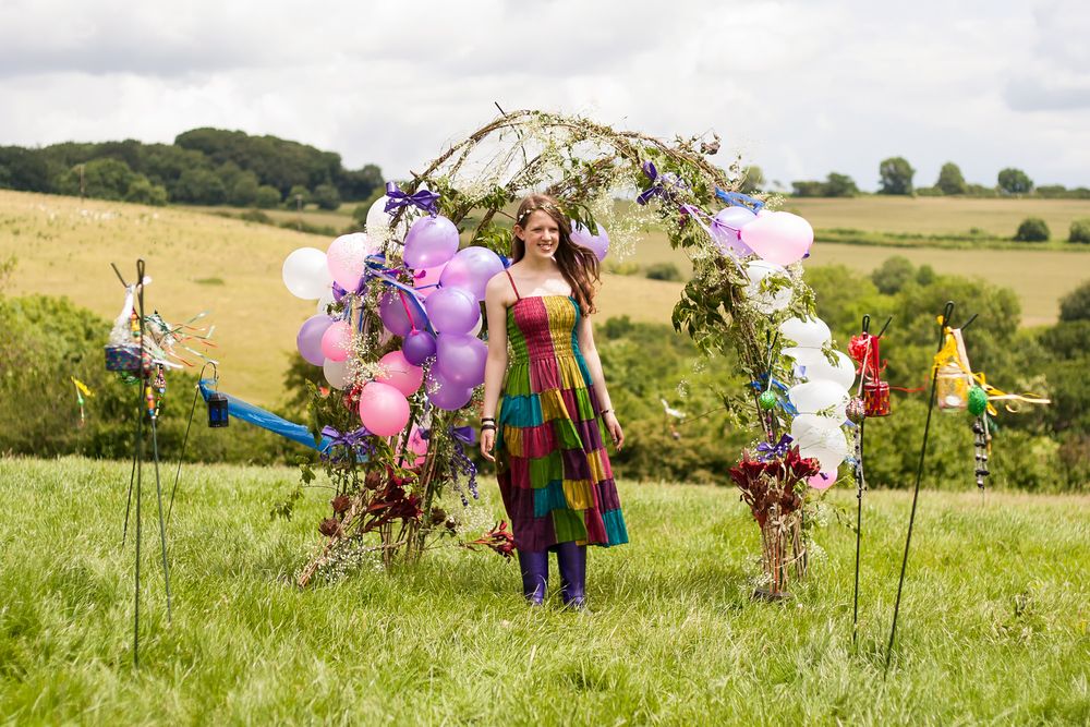 Portrait of a young lady in a colourful patchwork summer dress, posing under an arch of green boughs and balloons in a field.