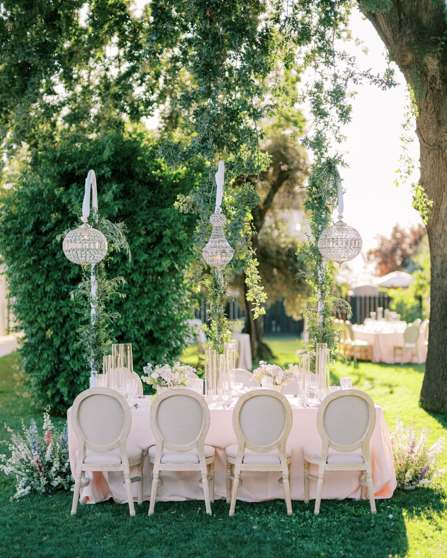 Elegant outdoor wedding table setup with white chairs and crystal chandeliers hanging from trees in a garden setting.