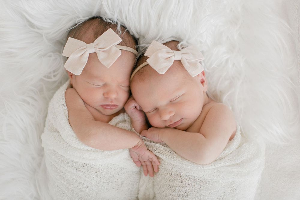 Two sleeping newborn babies wearing white bow headbands lie close together wrapped in soft white fabric.