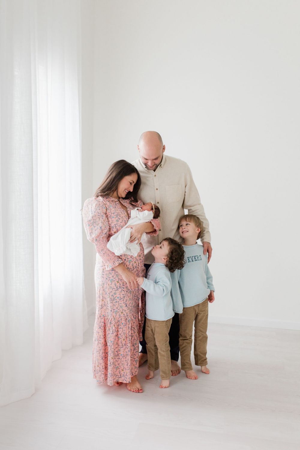 Family stands together in a bright white studio, wearing soft pastel colors against minimalist backdrop.