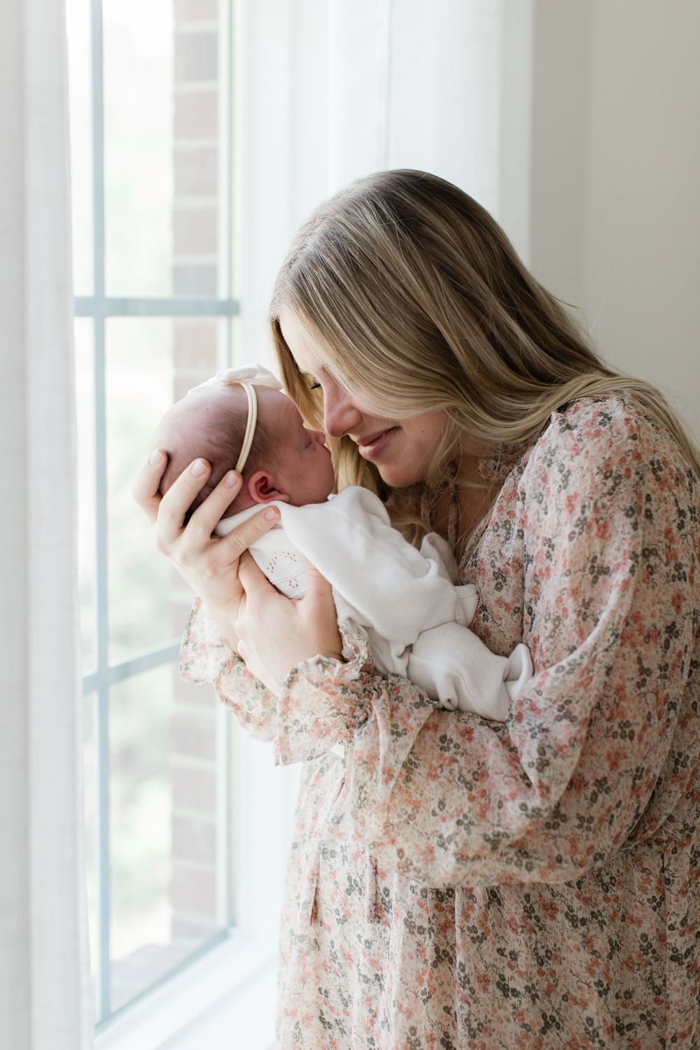 A tender moment by a bright window as a mother in a floral dress cradles and kisses her newborn baby.