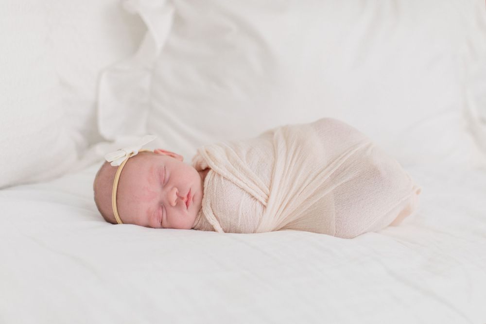 A newborn baby sleeps peacefully swaddled in white blankets on a soft white surface.