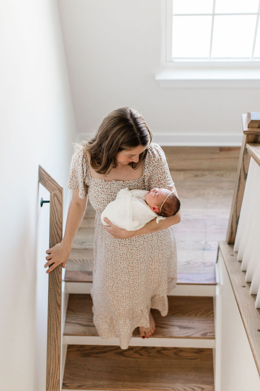 Woman in floral dress tenderly holds newborn baby while standing on wooden staircase in bright sunlit home.
