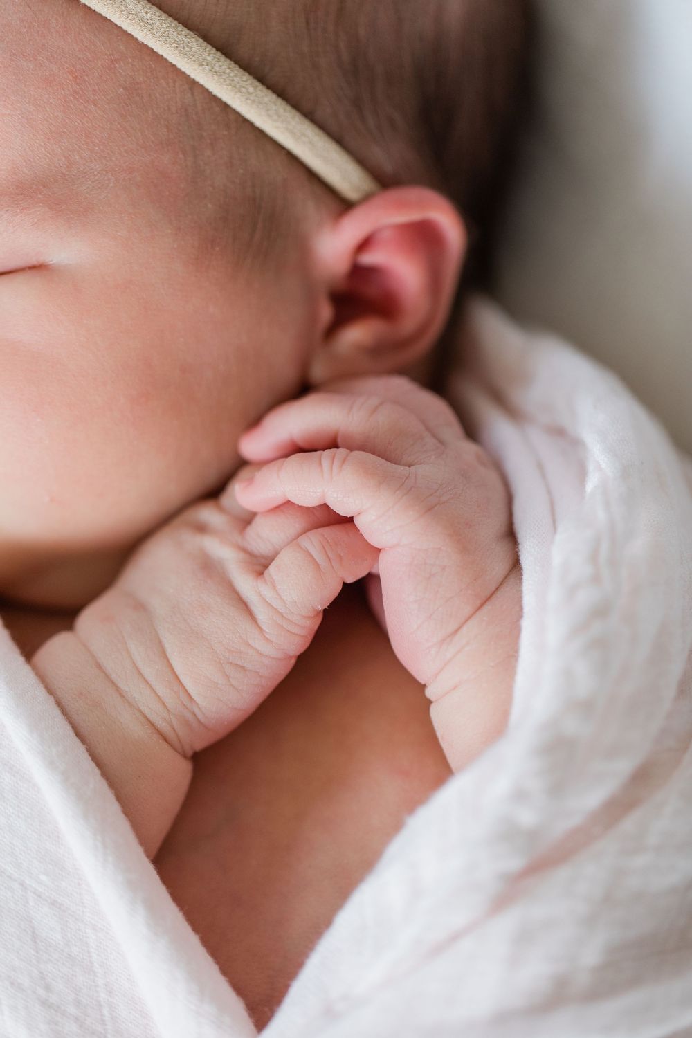 Close up of a tiny baby hand clasped near ear, wrapped in soft white blanket.