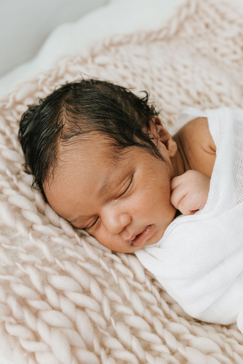 A sleeping newborn baby swaddled in white rests peacefully on a textured beige blanket.