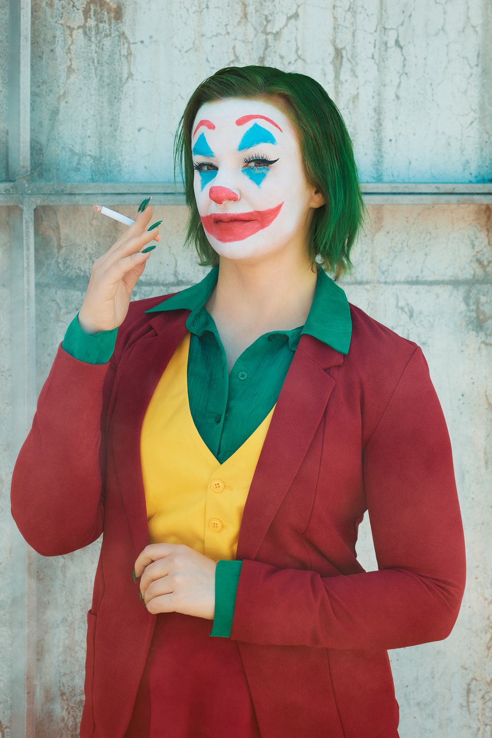 Cosplay of a clown character wearing green hair, white face paint, red suit jacket and yellow vest against concrete wall.