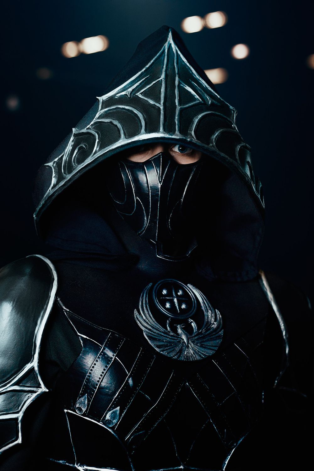 Dramatic cosplay photo of a dark armored figure wearing an ornate hooded helmet with glowing details in low light.