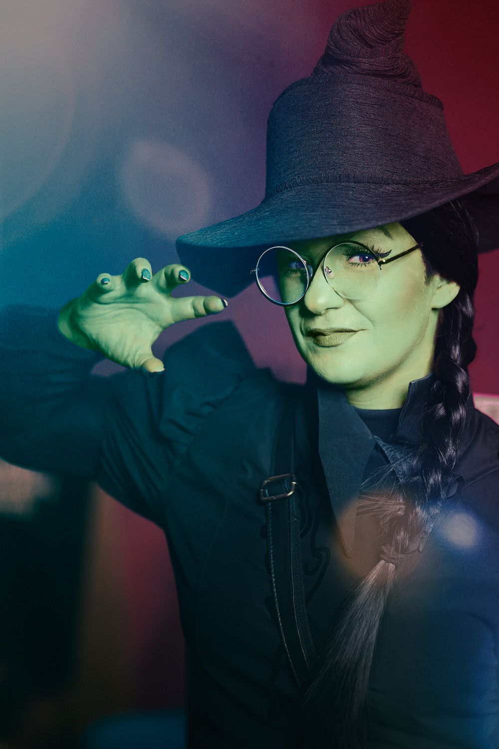Person in black witch costume and round glasses holds small toy frog in moody lighting.