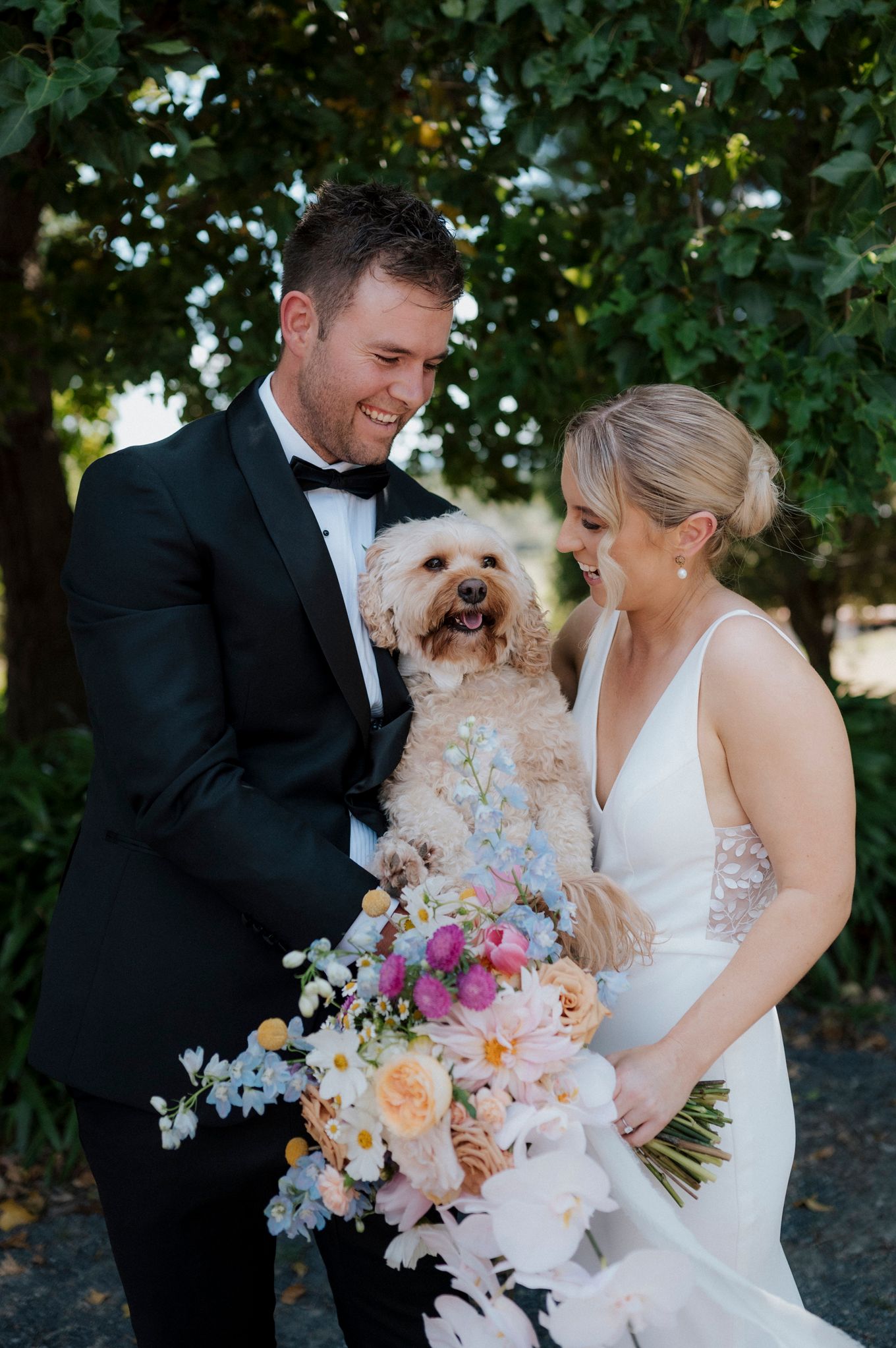 Wedding couple holds their small white dog while displaying colorful floral bouquet.