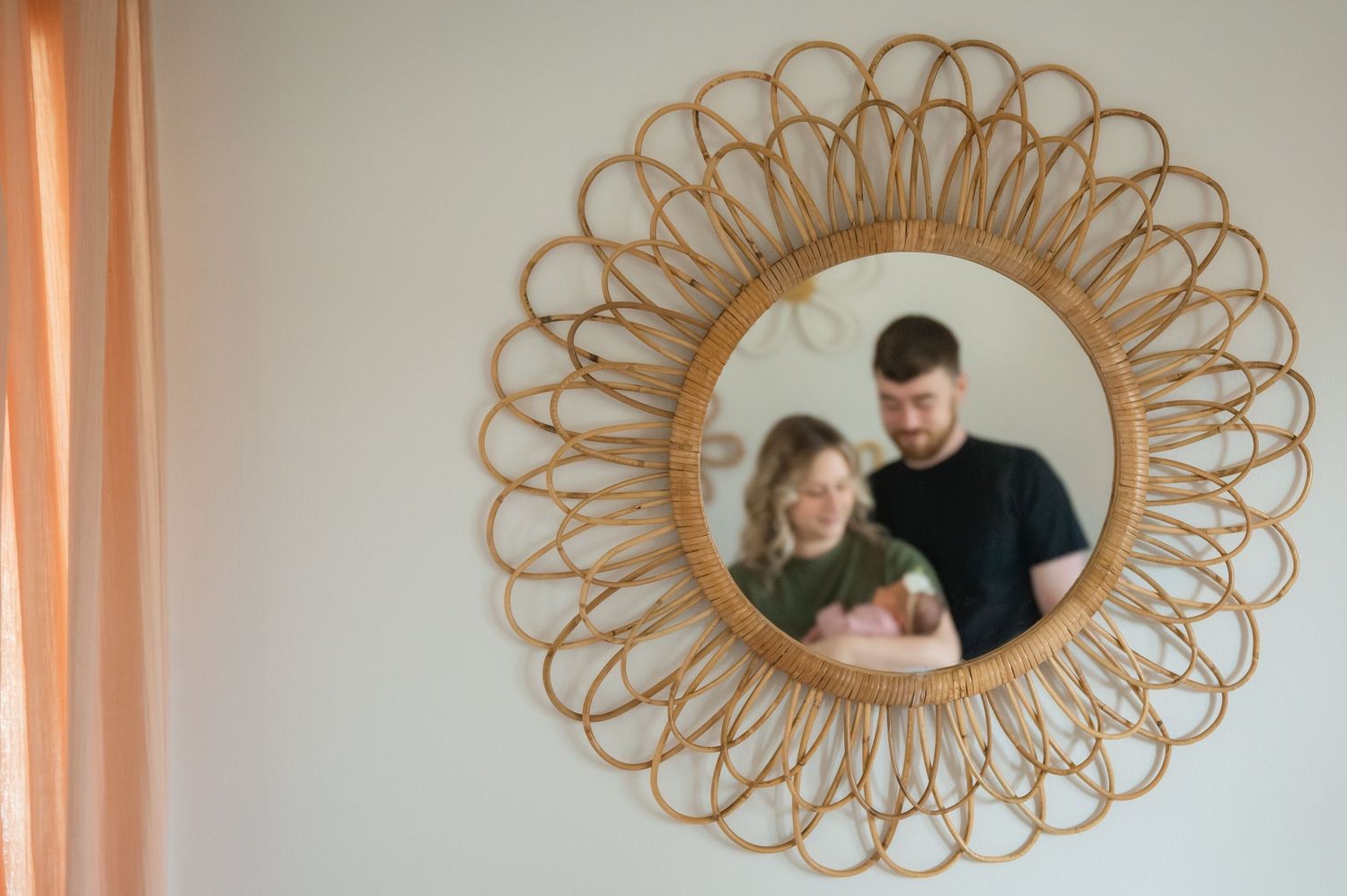 A couple stands behind a decorative rattan sunburst mirror frame while holding a small dog.