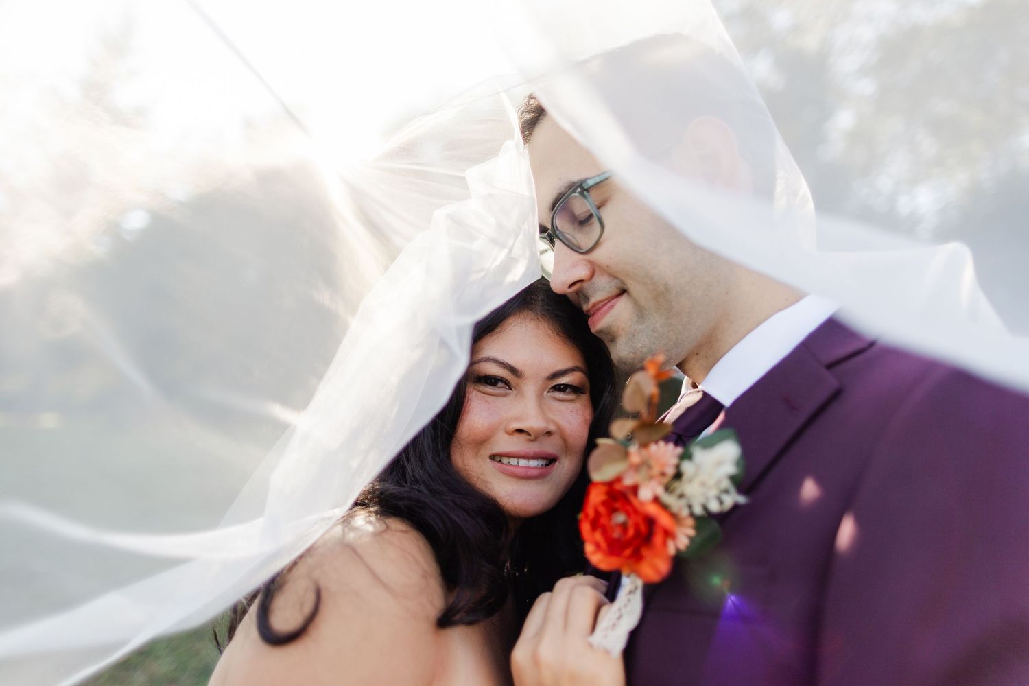 A couple shares a joyful moment under a flowing veil, with bright orange flowers on the groom's purple suit.