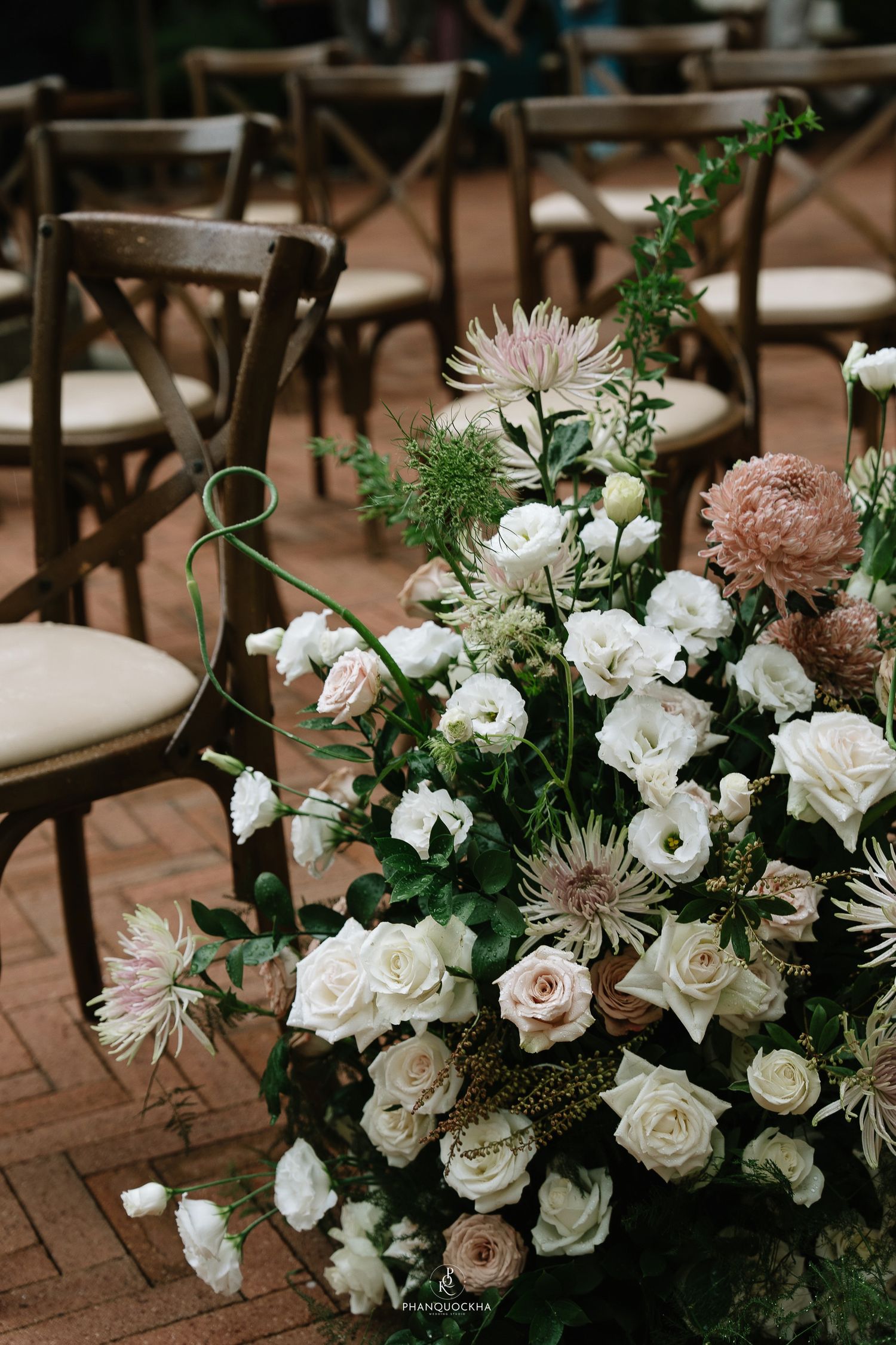 Lush wedding aisle floral arrangement with white roses, blush blooms, and greenery beside wooden chairs.