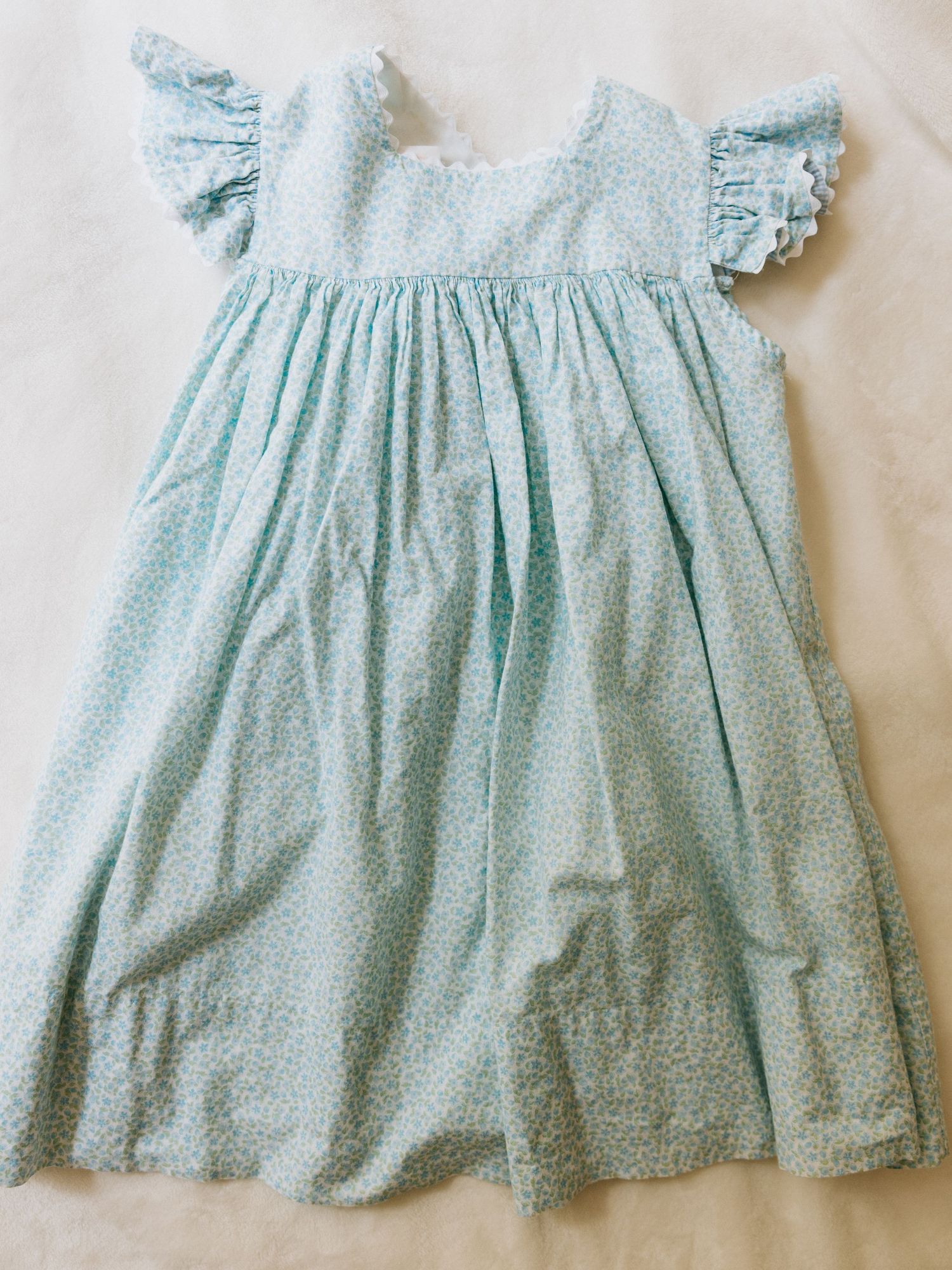 Formal infant outfit with beige vest, white shirt, light blue bow tie, and matching shorts hanging on wooden ladder shelf.