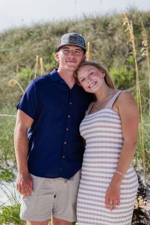 A couple poses together on a beach, wearing casual summer attire with sea grass in the background.