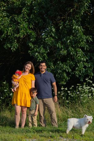Family and their white dog pose together in a natural setting with tall grass and trees.