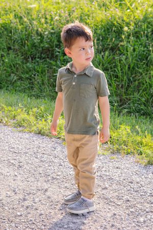 A small child in khaki pants and olive polo shirt stands on a gravel path.