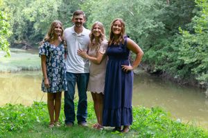 A group poses together outdoors on a grassy slope near a pond surrounded by lush summer greenery.