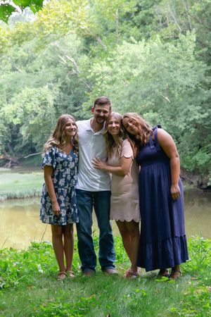 Friends gather for a casual outdoor photo on a grassy bank with trees and water in the background.