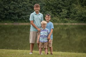 Three siblings in coordinating blue and khaki outfits stand together by a lake.