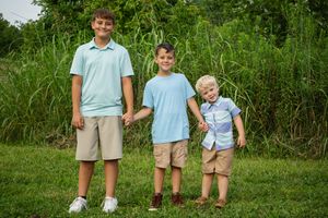 Three brothers in matching light blue polos and khaki shorts stand in height order on grass.