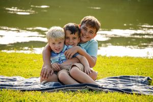 Three siblings share a warm embrace while sitting on a blanket near a lake.