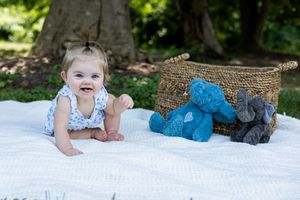 Baby lying on white blanket outdoors next to woven basket and blue plush toys.