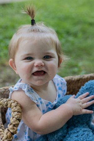 Smiling baby with tiny ponytail wearing blue floral dress next to woven basket.