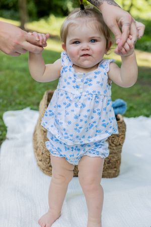 Baby in blue floral outfit standing with support on white blanket outdoors.