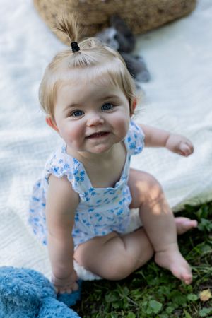 Baby with hair tuft in blue floral outfit sitting on white blanket outdoors.
