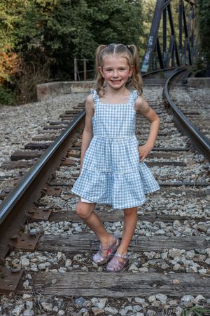Young person in blue gingham dress poses playfully on railroad tracks surrounded by rocks.